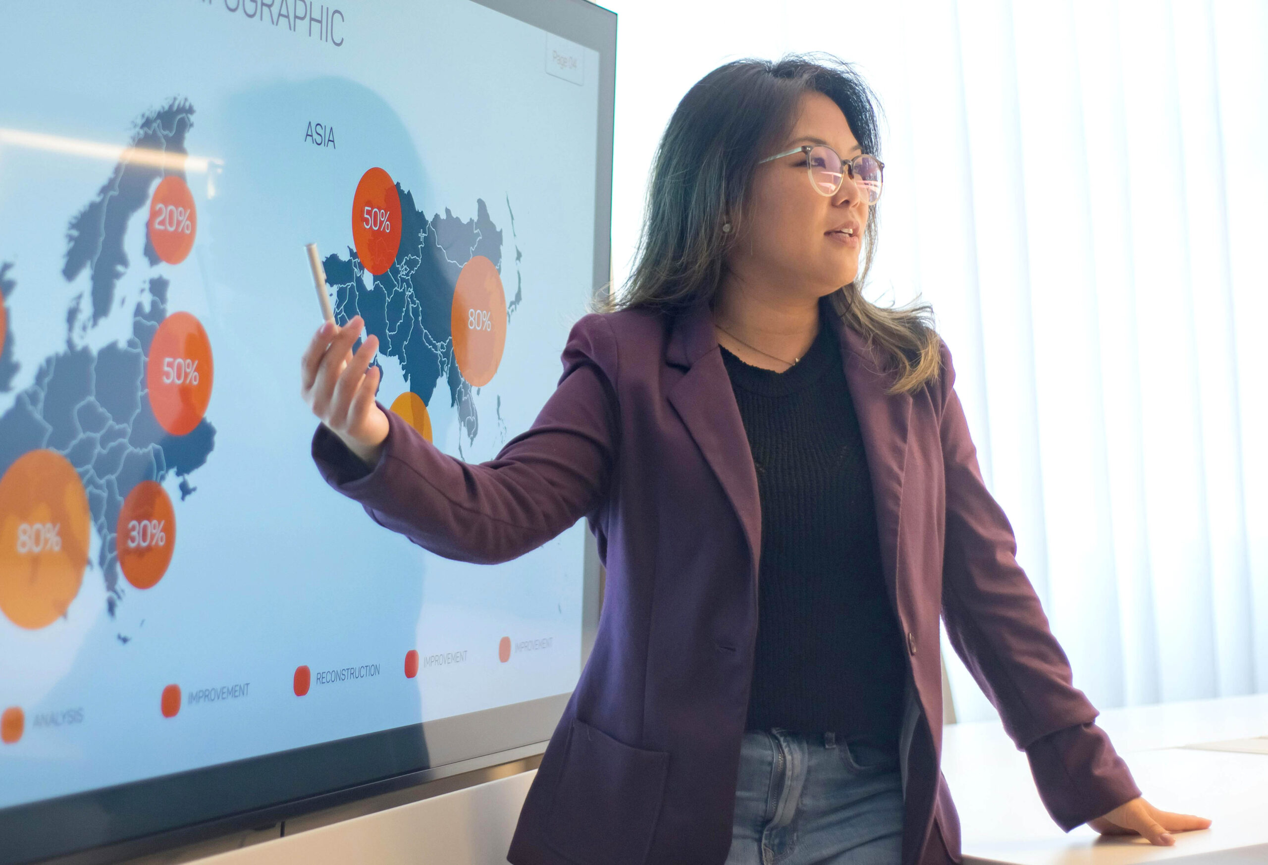 a younger professional woman dressed in businss casual attire is smiling as she stands next to a flipboard on an easel. A young man is sitting on a green velvet couch on the left hand side of the scene. A lady is sitting in a chair facing the presenter on the right hand of the scene.